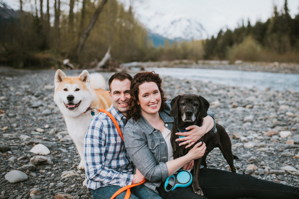 Couple hugging their dogs and smiling at the camera by riverbed