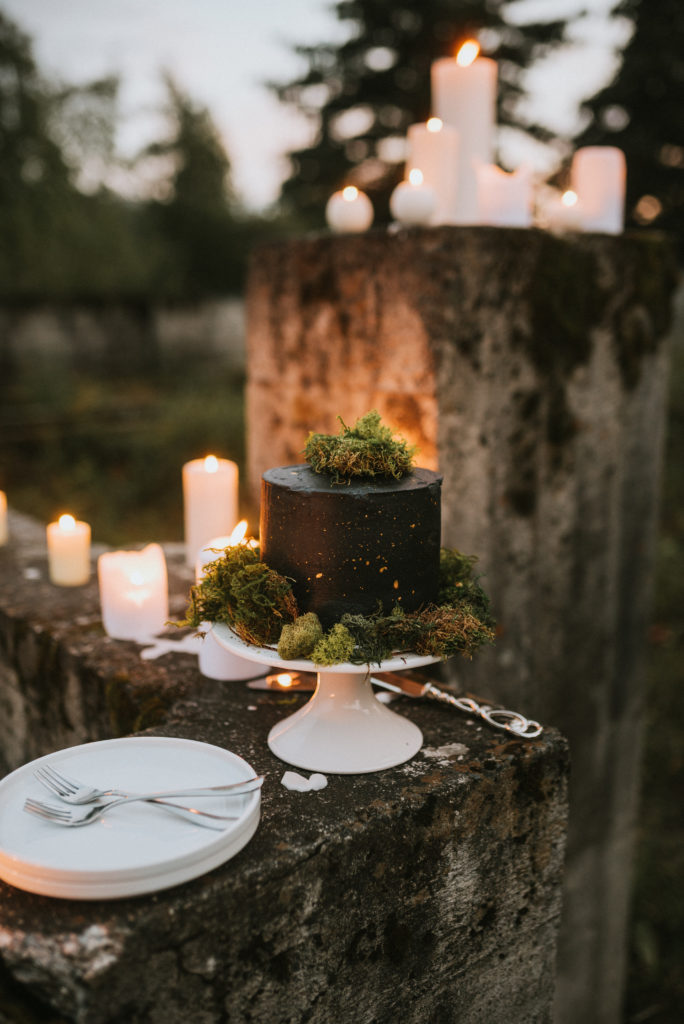 Black gold splattered cake with moss and candlelight against concrete backdrop