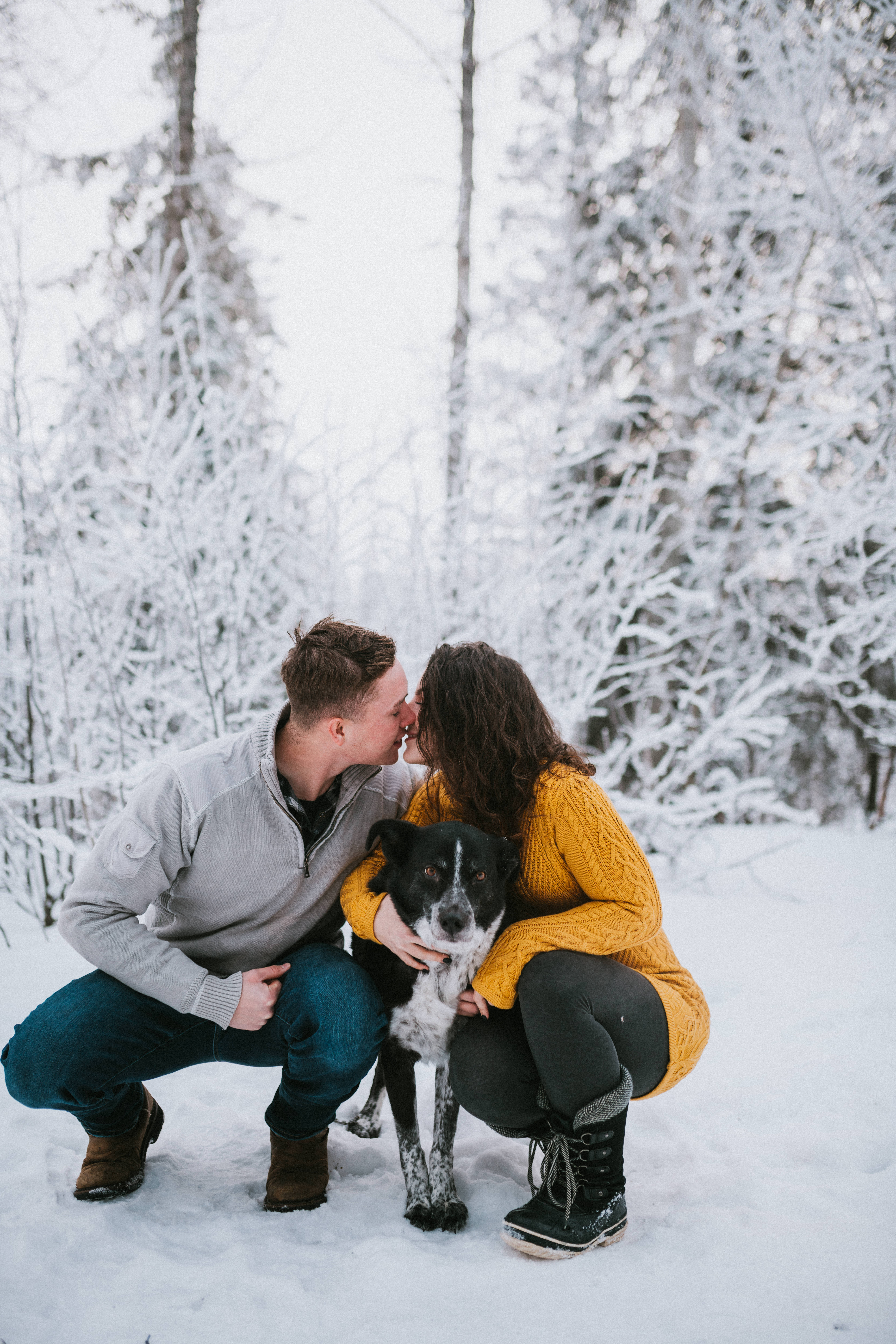 Couple crouching in snow with their dog and kissing during engagement session