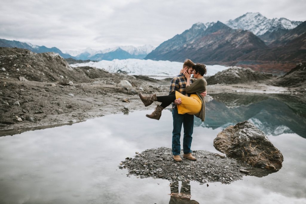 A romantic photo of a glacier lake at Matanuska Glacier Park as a couple stands in the middle and he is holding her bridal style.