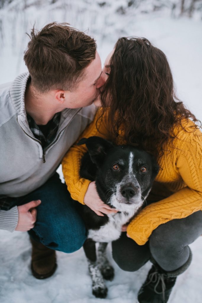 Couple kissing each other whiles snuggling their dog during their winter engagement session.