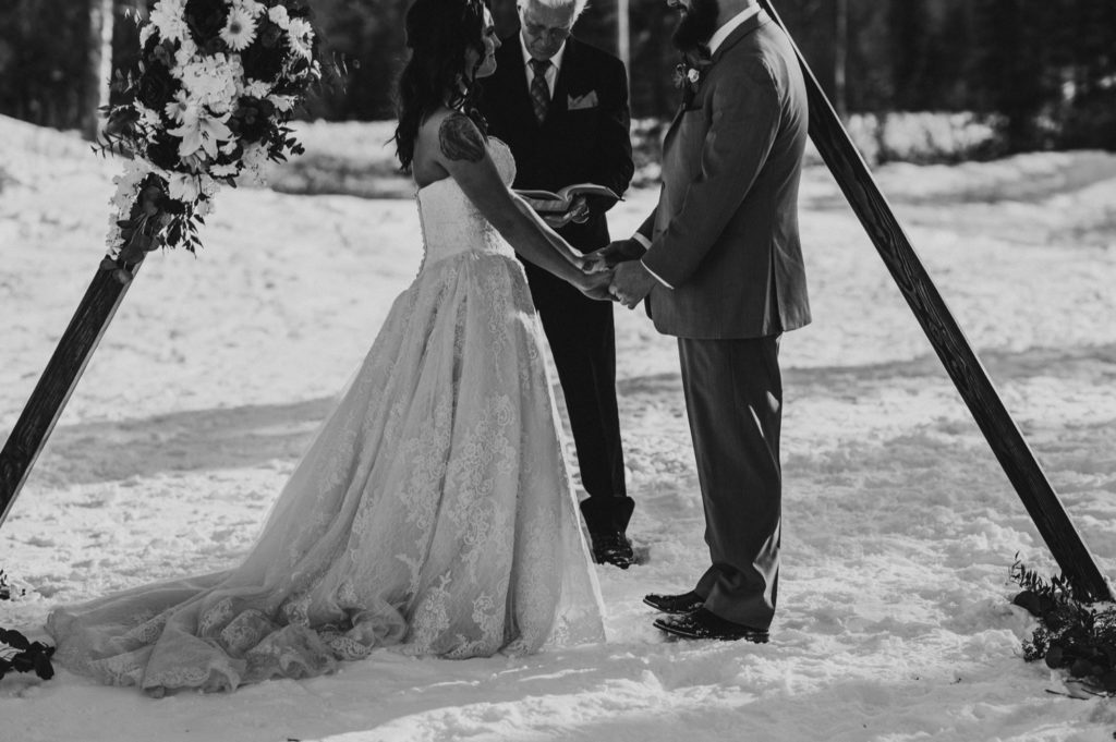 Black and white detail photo of bride and groom holding hands during ceremony at Alaska winter wedding