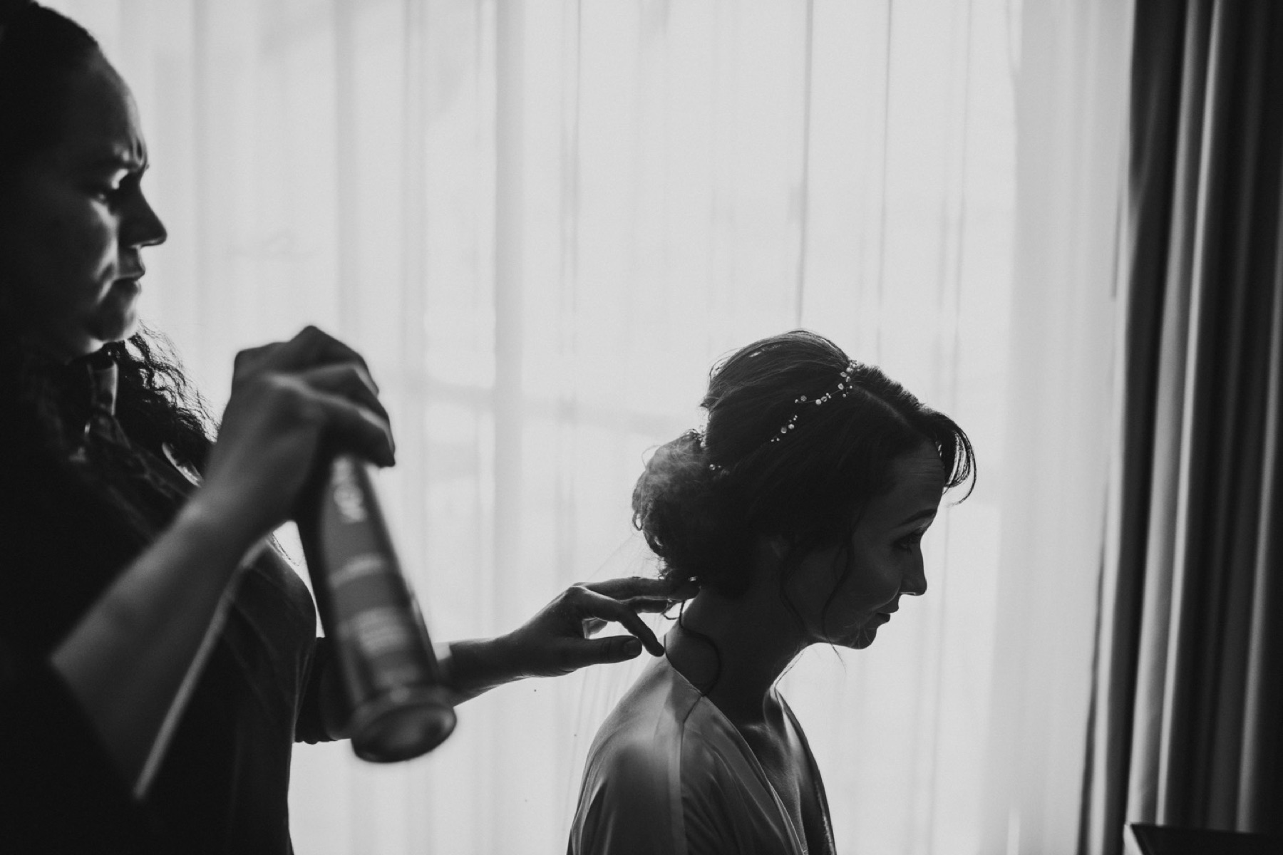 Black and white photo of a bride getting the last touches of hair spray in front of a window