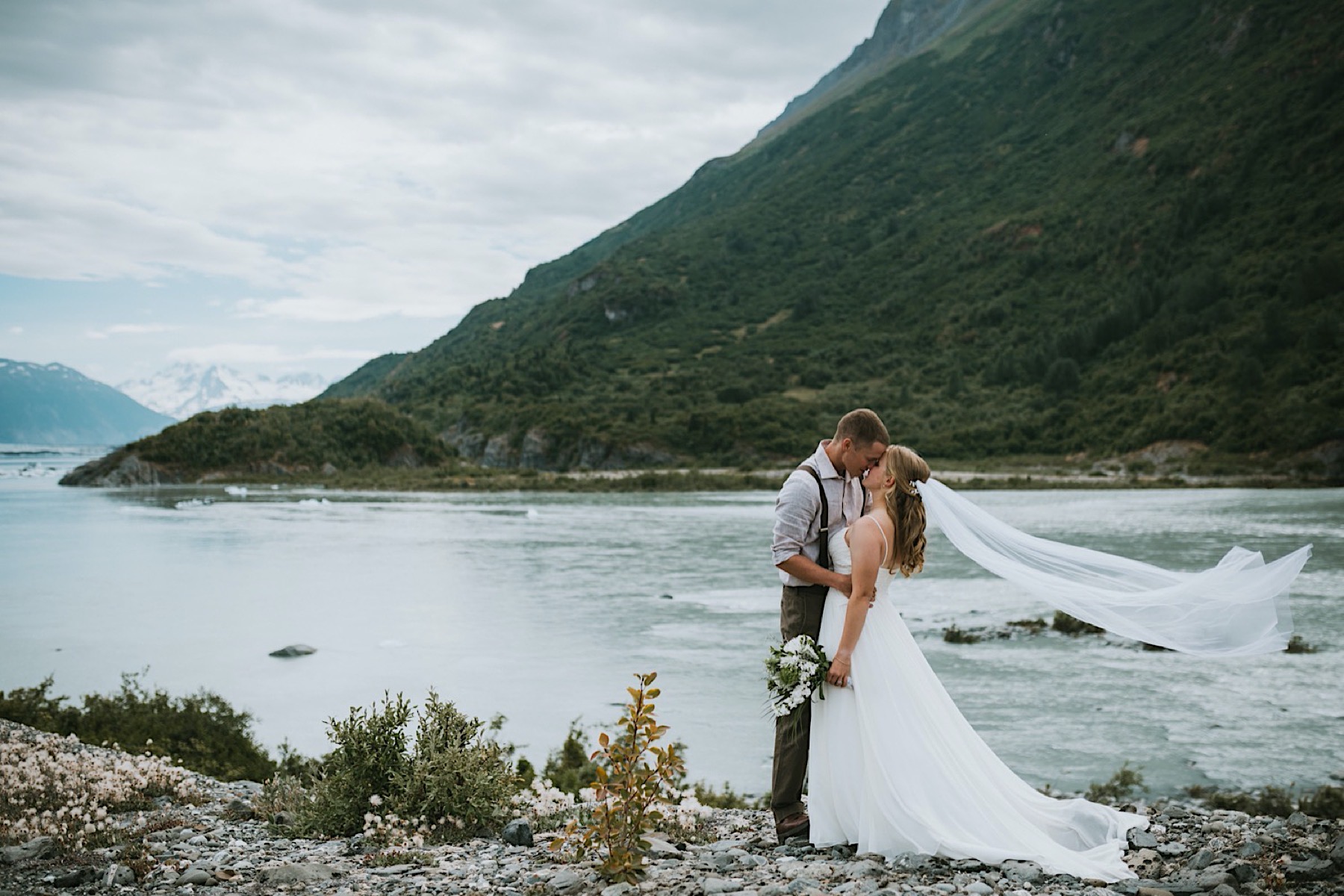 Bride and groom kissing in front of Knik River as her veil blows in the wind