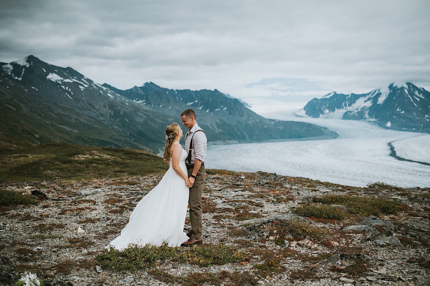 Bride and groom holding hands in front of Knik Glacier smiling at each other during Alaska Destination wedding