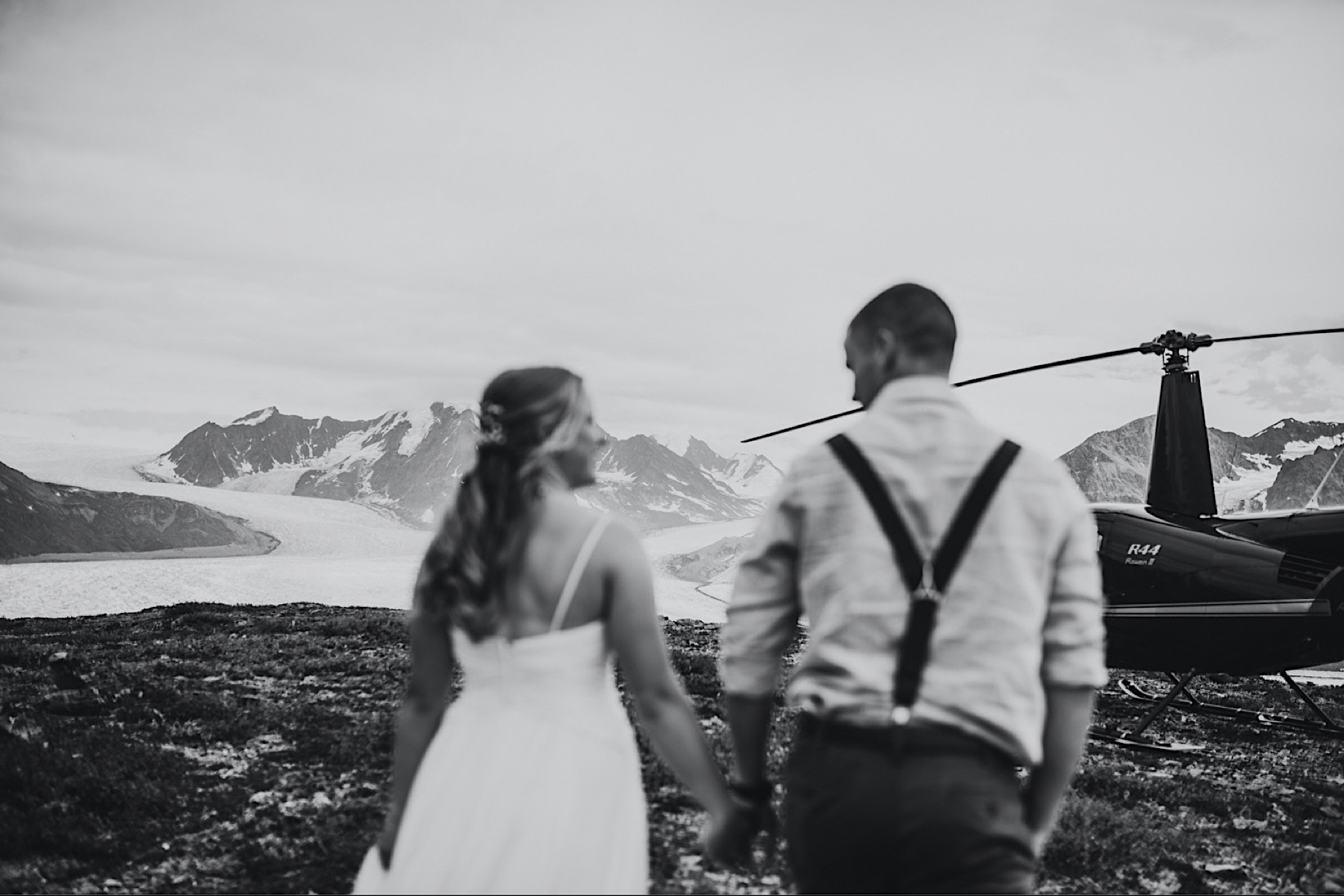 Black and white photo of bride and groom walking towards helicopter with mountains in focus in front of them