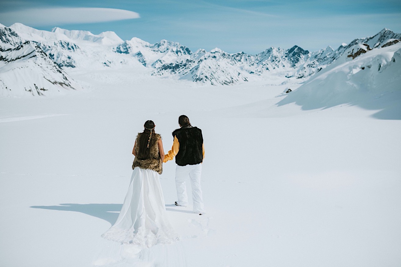 Bride and groom holding hands and walking across a snow packed glacier with bright blue skies and mountain peaks