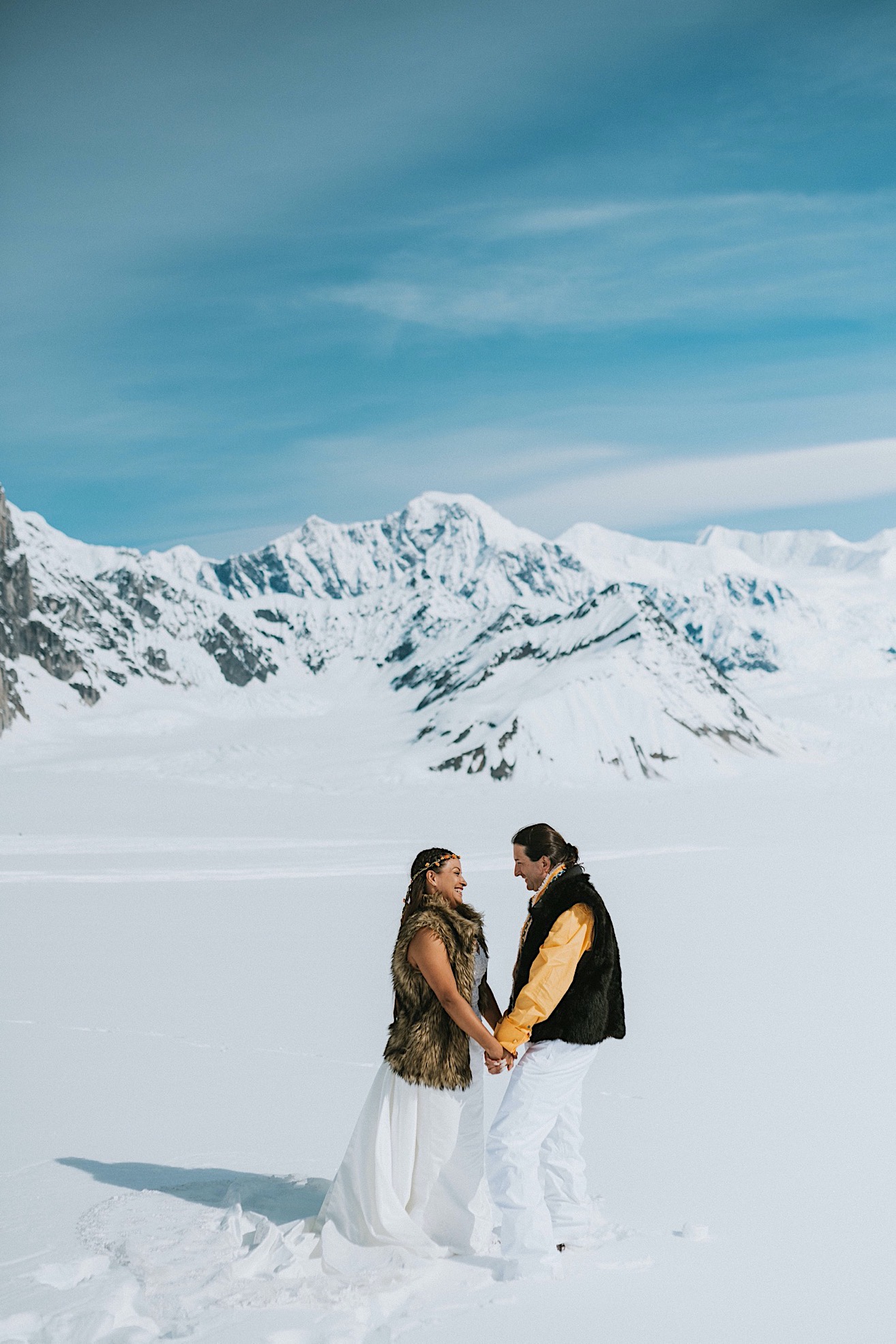 Bride and groom in fur vets holding hands looking at each other on a glacier