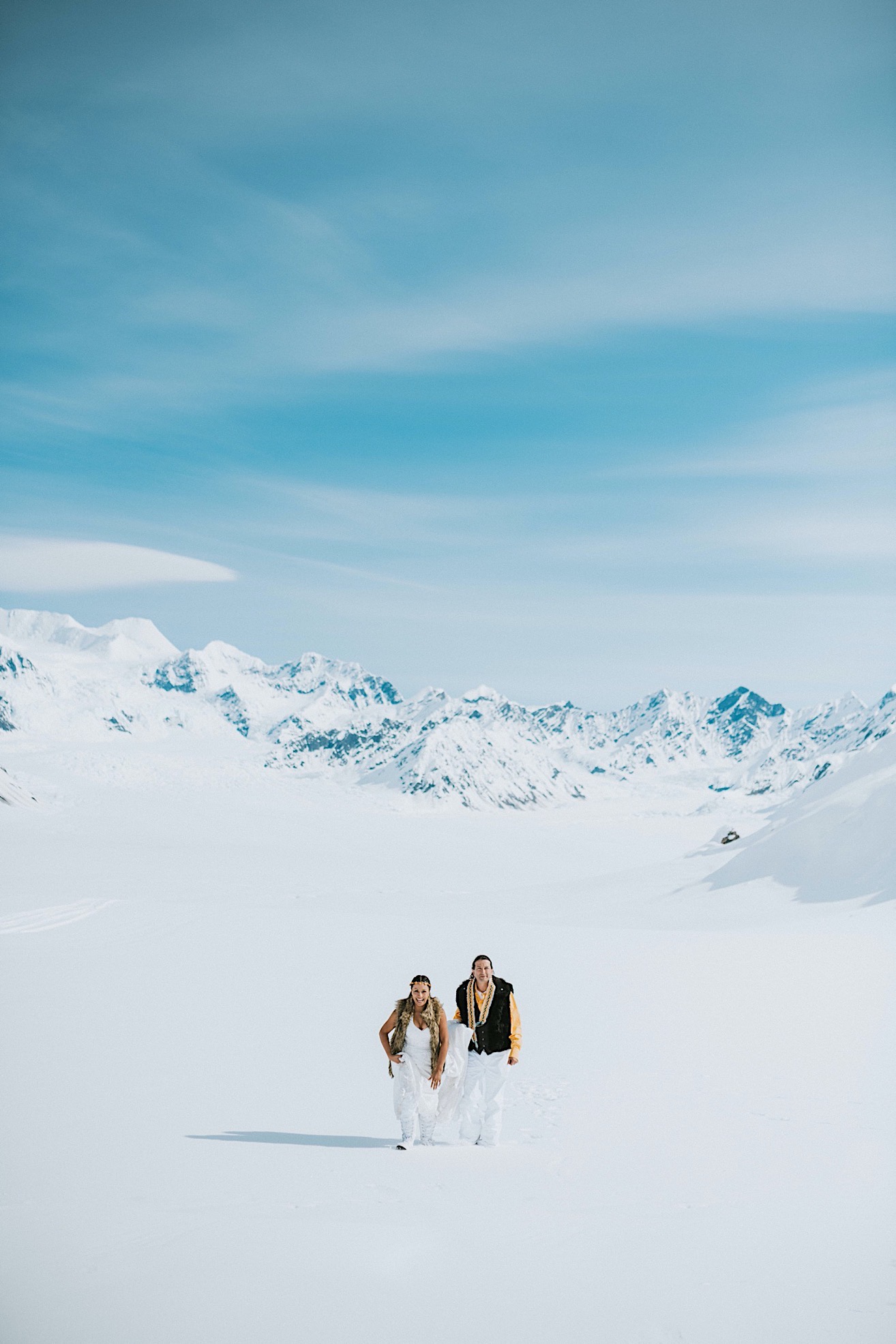 Bride and groom wearing fur vests walking across pristine snow on Ruth Glacier after their glacier elopement