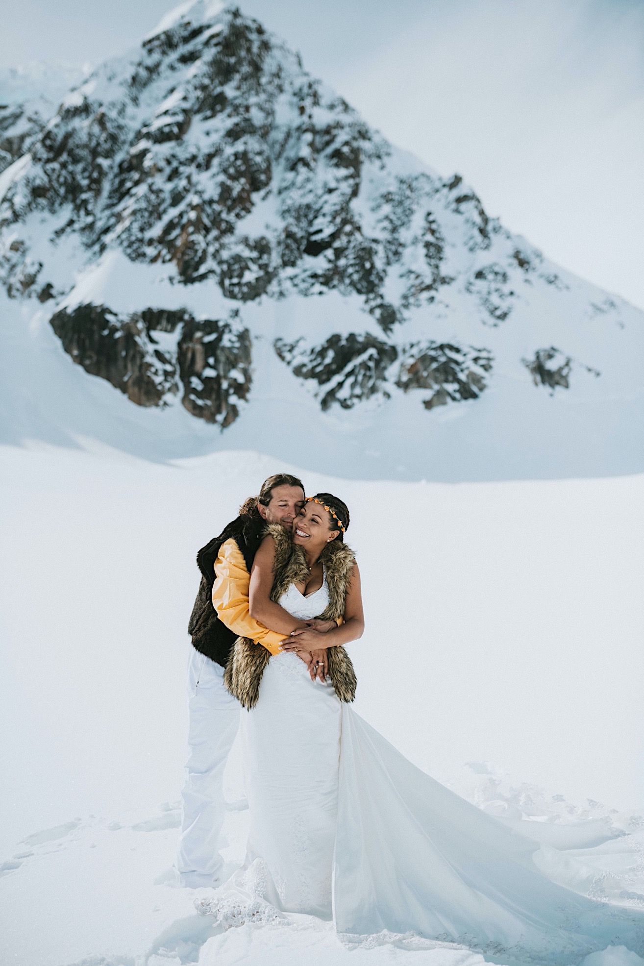 Groom bear hugging his bride and kissing her in front of a mountain peak on Ruth Glacier