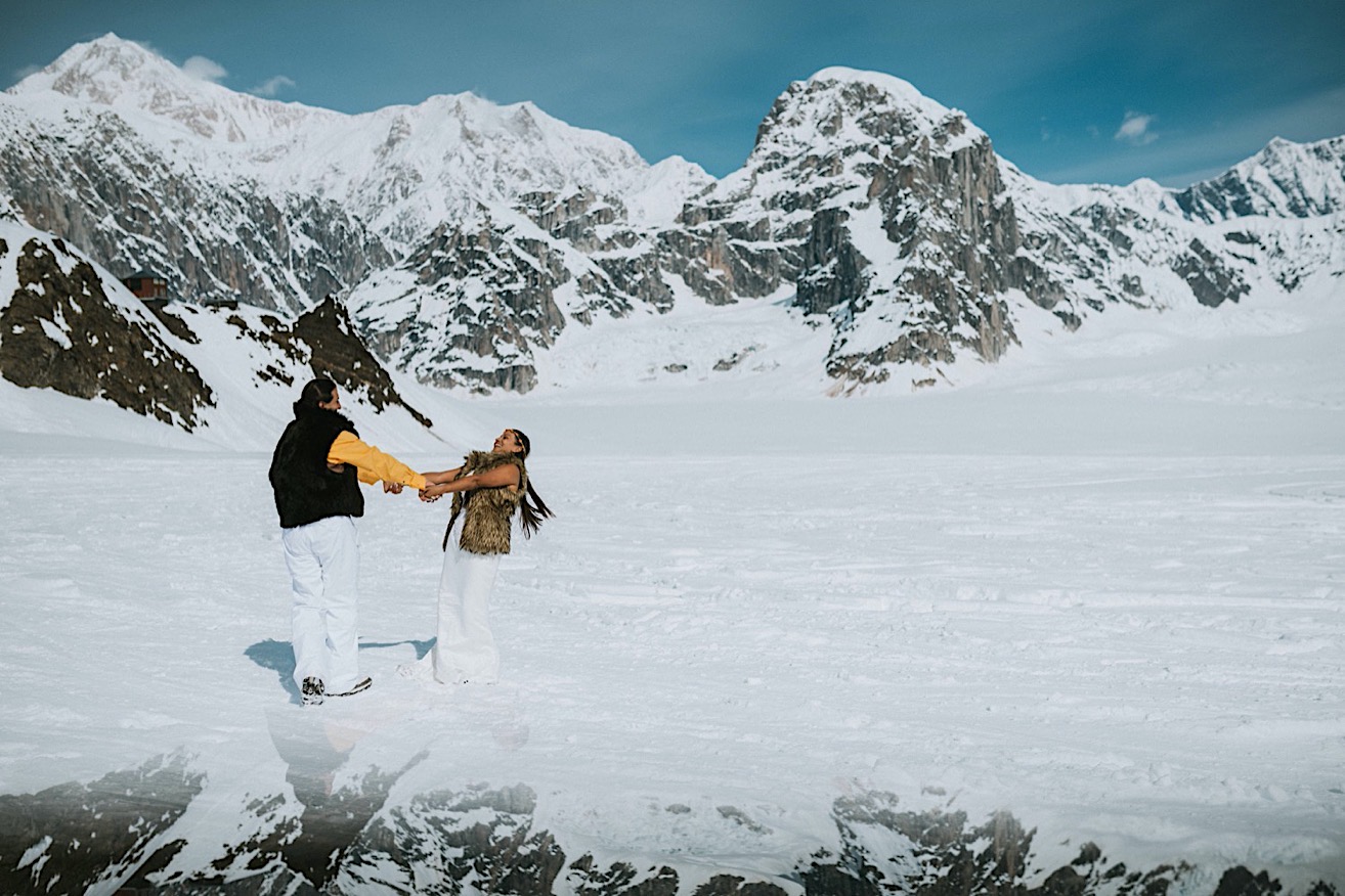 Bride and groom wearing fur vests holding hands and spinning in the snow with the mountains reflected around them