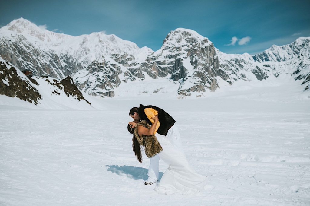 Bride and groom wearing fur vests dip kissing in front of Denali