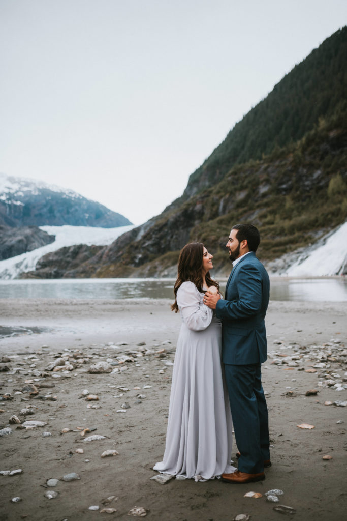 Couple holding hands and looking at each other in front of Mendenhall Glacier and Nugget Falls in Juneau during Engagement photos in Alaska coastal town