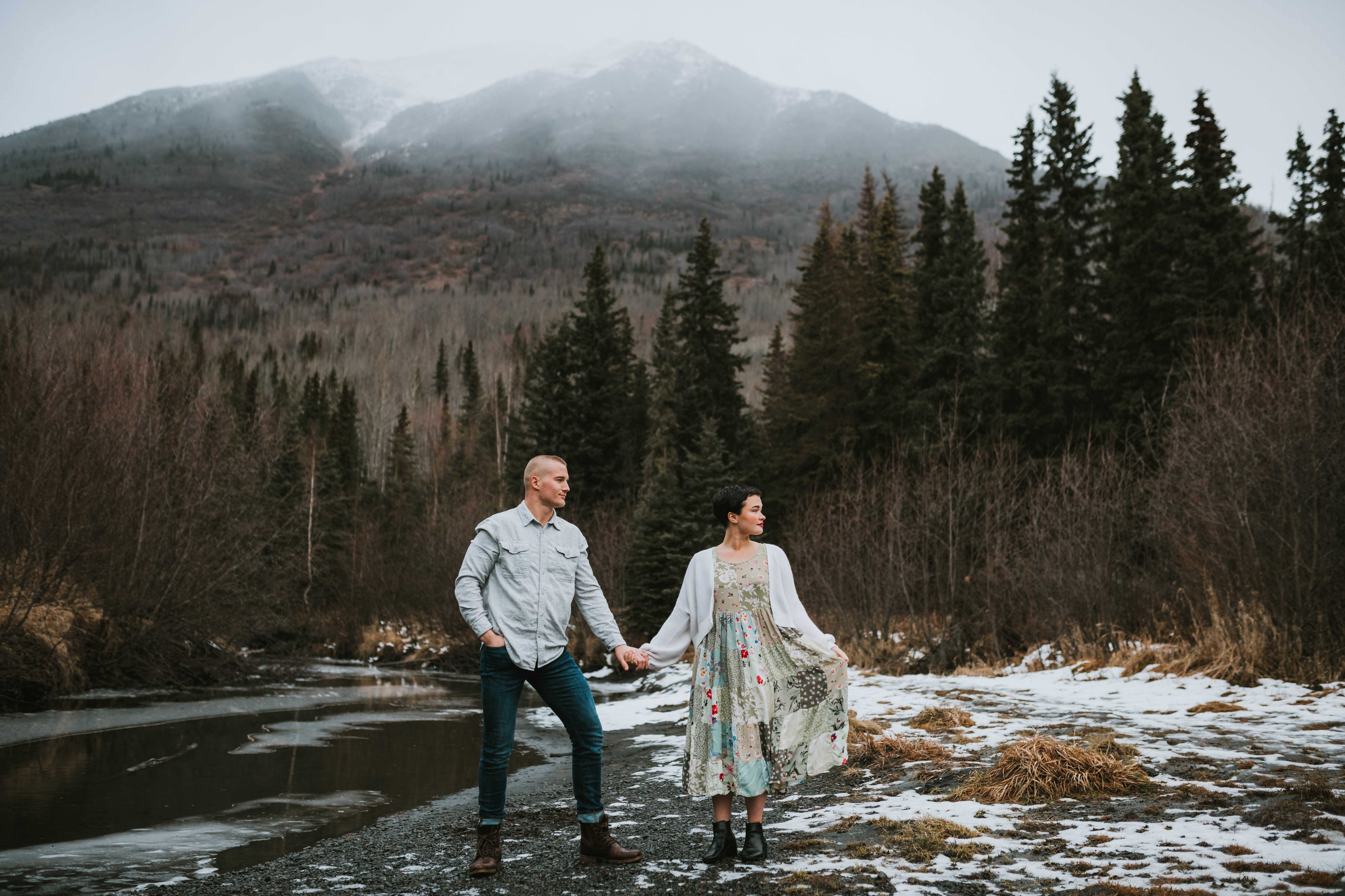Couple posed looking into the distance while she lifts the corner of her dress against the mountains of North Fork Eagle River