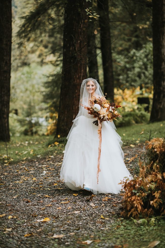 Bride wearing tulle dress walking down gravel trail in the woods to her first look at Raven Glacier Lodge