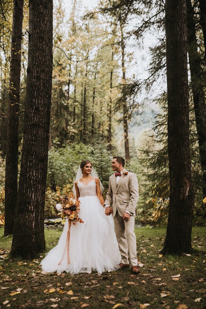 Bride and groom standing side by side looking at each other smiling amidst fall colored foliage and tress at Raven Glacier Lodge