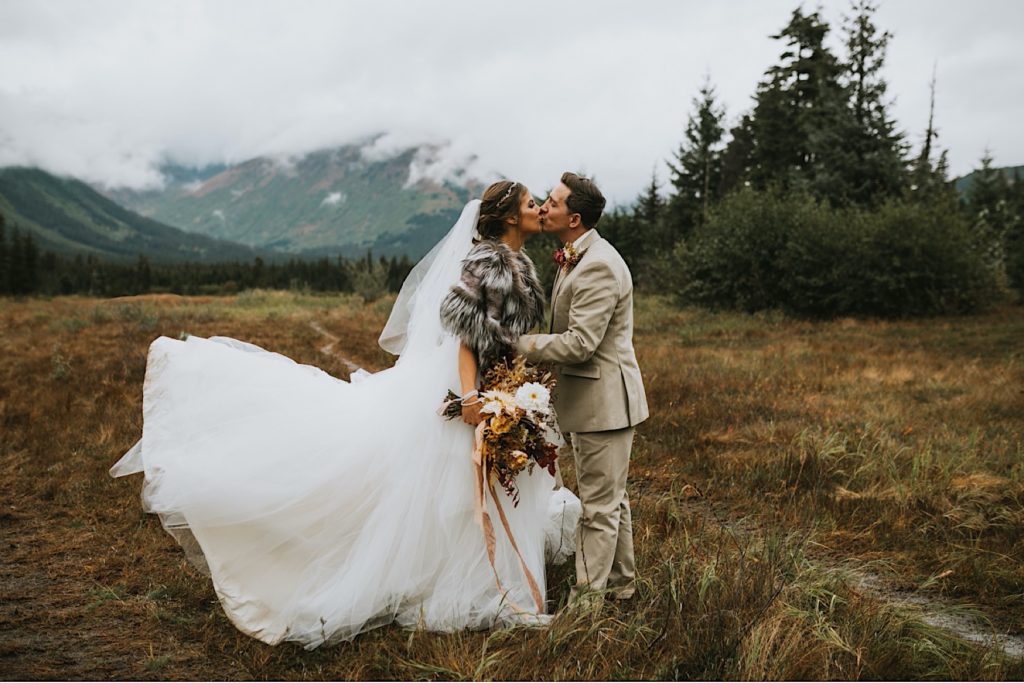Romantic images of bride and groom kissing in front of Girdwood mountains with her dress being tossed in the air