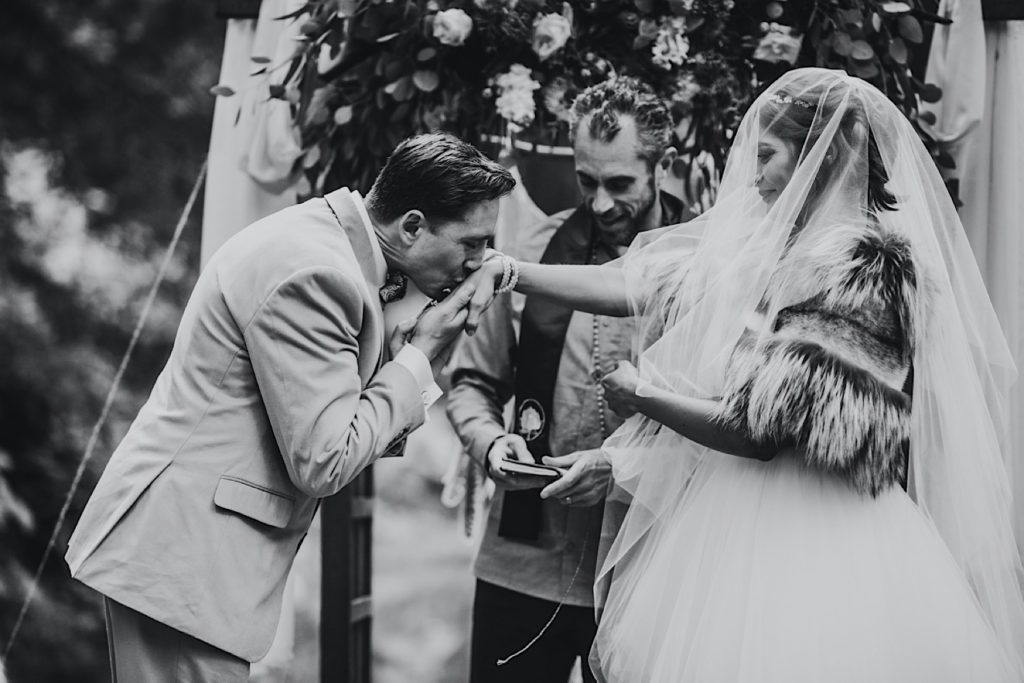 Black and white image of groom kissing his brides hand during ceremony