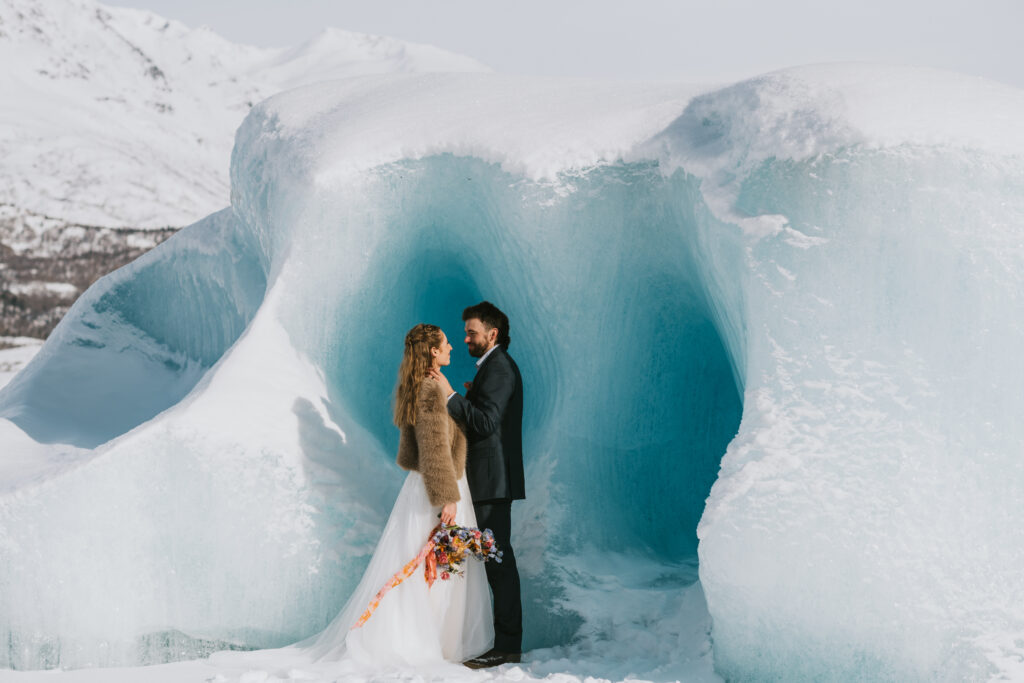 couple standing in front of a blue iceberg in their wedding attire in alaska