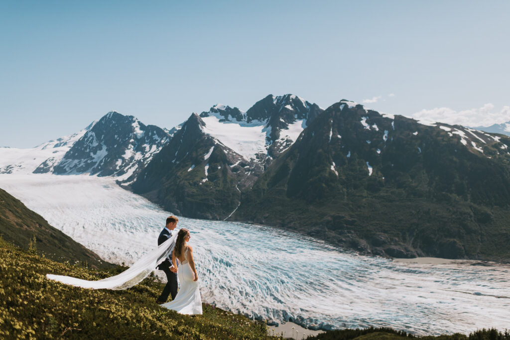 A man and woman elope in Alaska, walking on a mountain in their wedding dress.