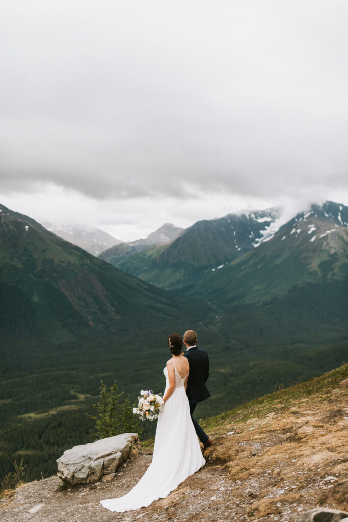 A bride and groom elope in Alaska, standing on top of a cliff overlooking the mountains.