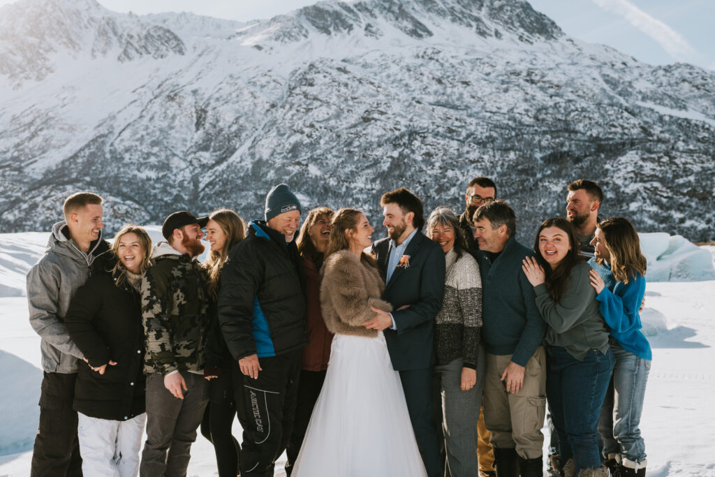 Exuberant wedding party gathered around the bride and groom, sharing laughter and close embraces on a snowy Alaskan landscape with a rugged mountain backdrop during an elopement.