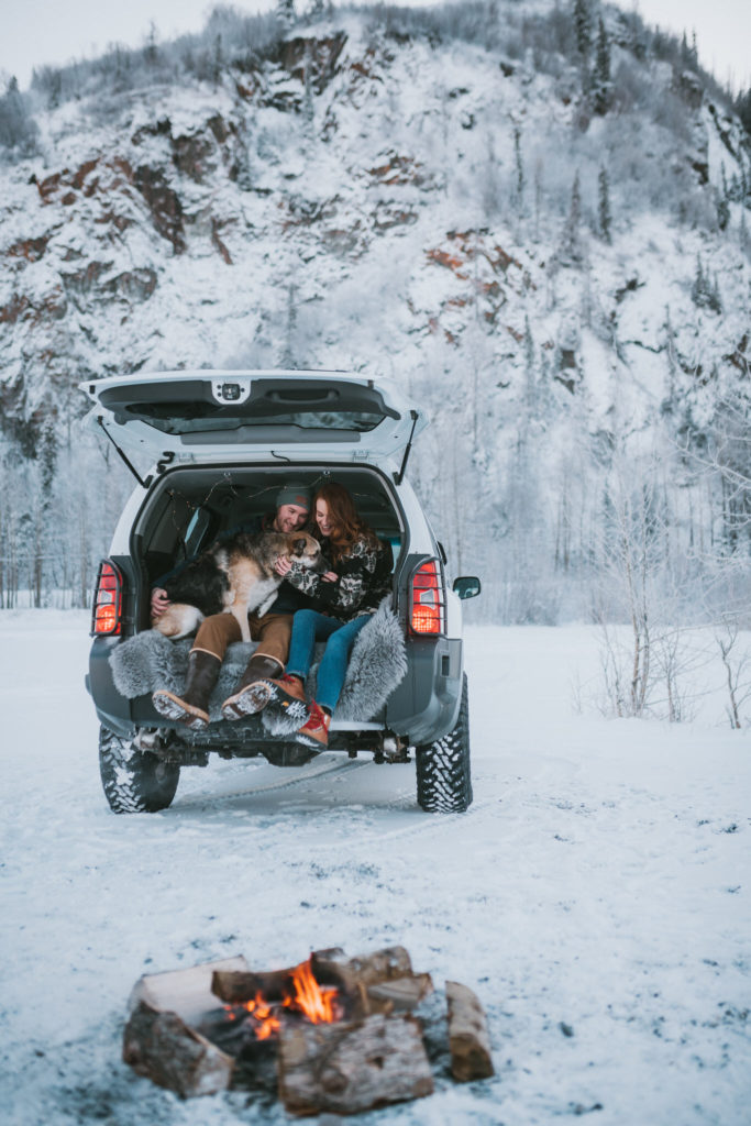 Couple sitting in the back of an xterra with campfire and german shepherd in the winter