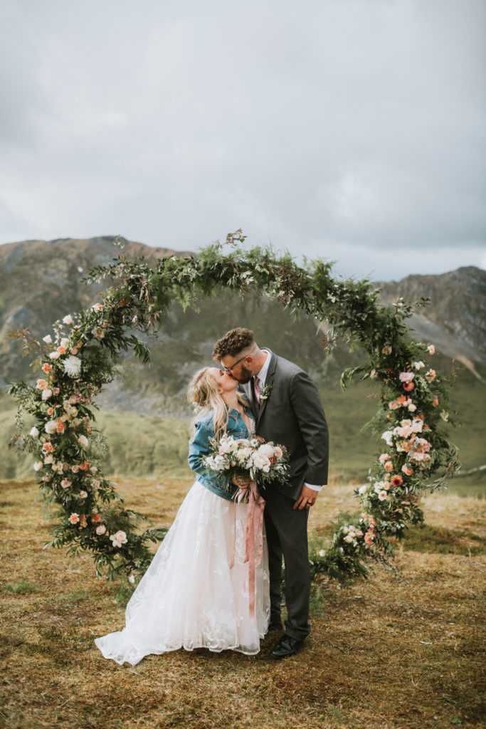 Couple kissing in middle of round floral arch in the mountains