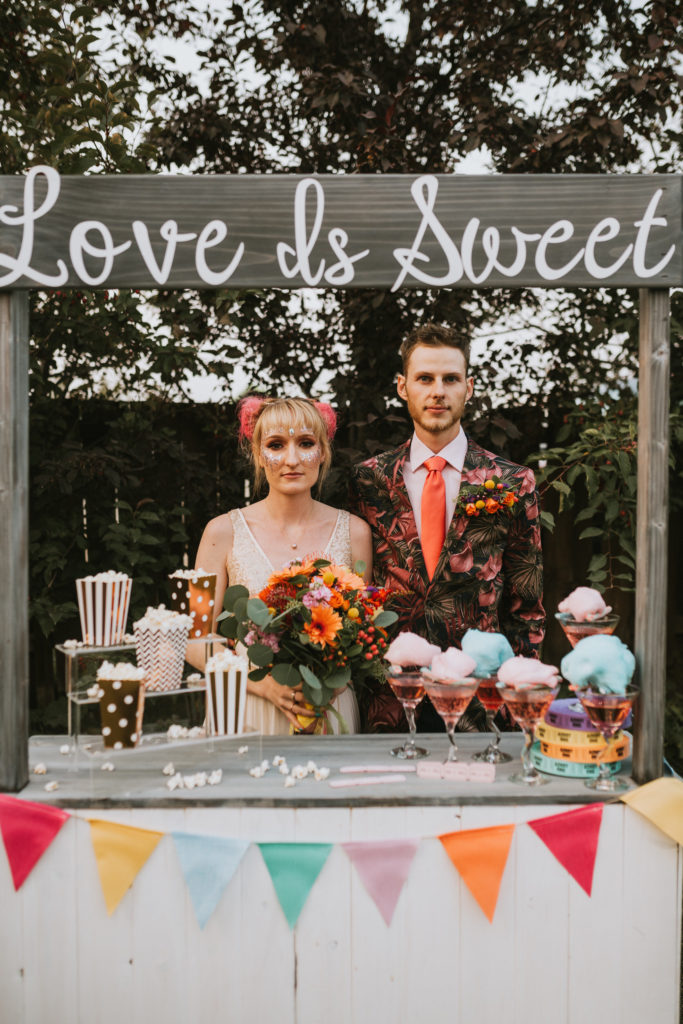 Colorful bride and groom standing at fair drink stand with colorful flowers and cotton candy cocktails