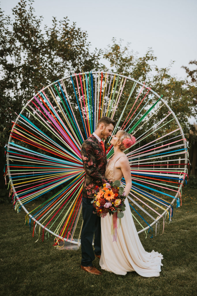 Bride and groom standing with colorful flowers in front of rainbow circle wedding arch for their ceremony
