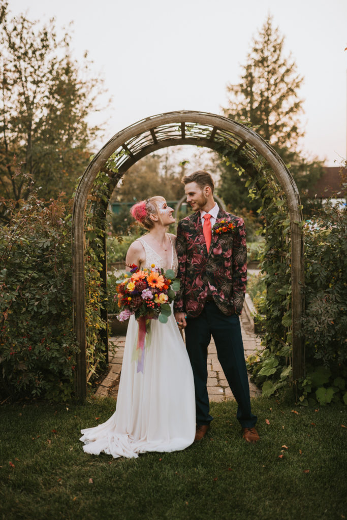 Colorfully dressed bride and groom standing in front of a garden arch smiling at each other
