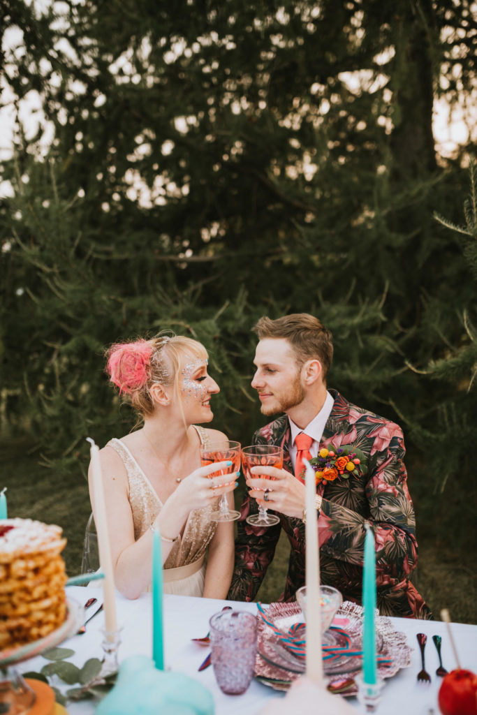 Bride and groom dressed in colorful wedding attire with florals and sparkles cheersing their cotton candy cocktail