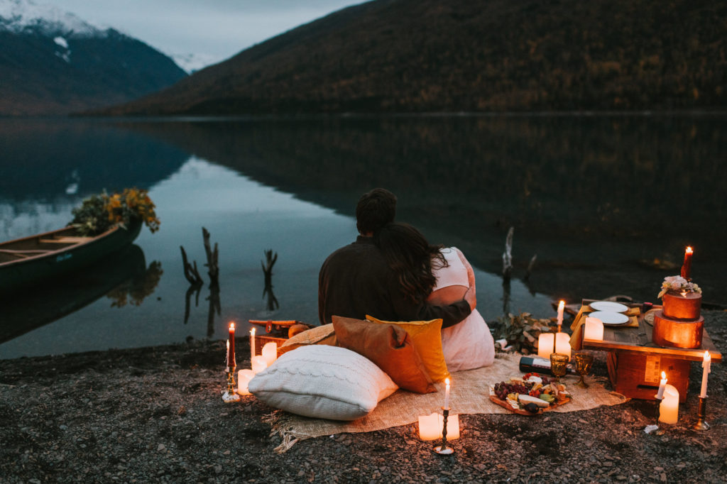 Couples sitting with their heads together at a lakeside candlelit picnic with mountain landscapes