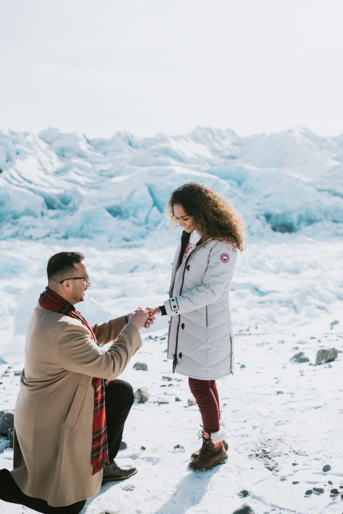 photo of a couple on a glacier and he is putting a ring on her finger after a surprise proposal
