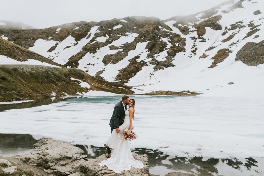 wedding couple standing at the edge of a mostly frozen summit lake in hatcher pass alaska