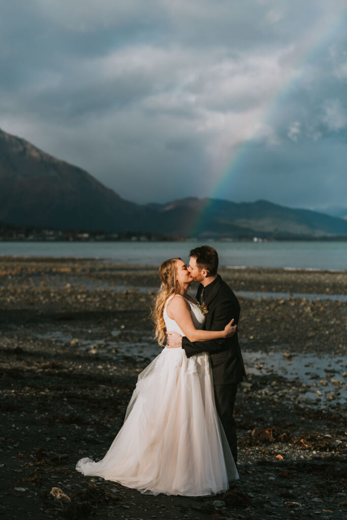 wedding couple kissing under a rainbow on a beach in seward alaska on a sunny day