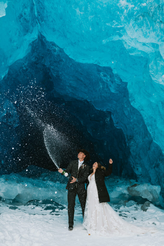 A man and woman standing under a blue ice cave spraying champagne