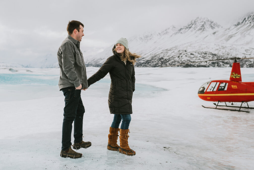 A couple holding hands on a frozen landscape with snow-covered mountains in the background and a red helicopter parked nearby, posing for their engagement photos.