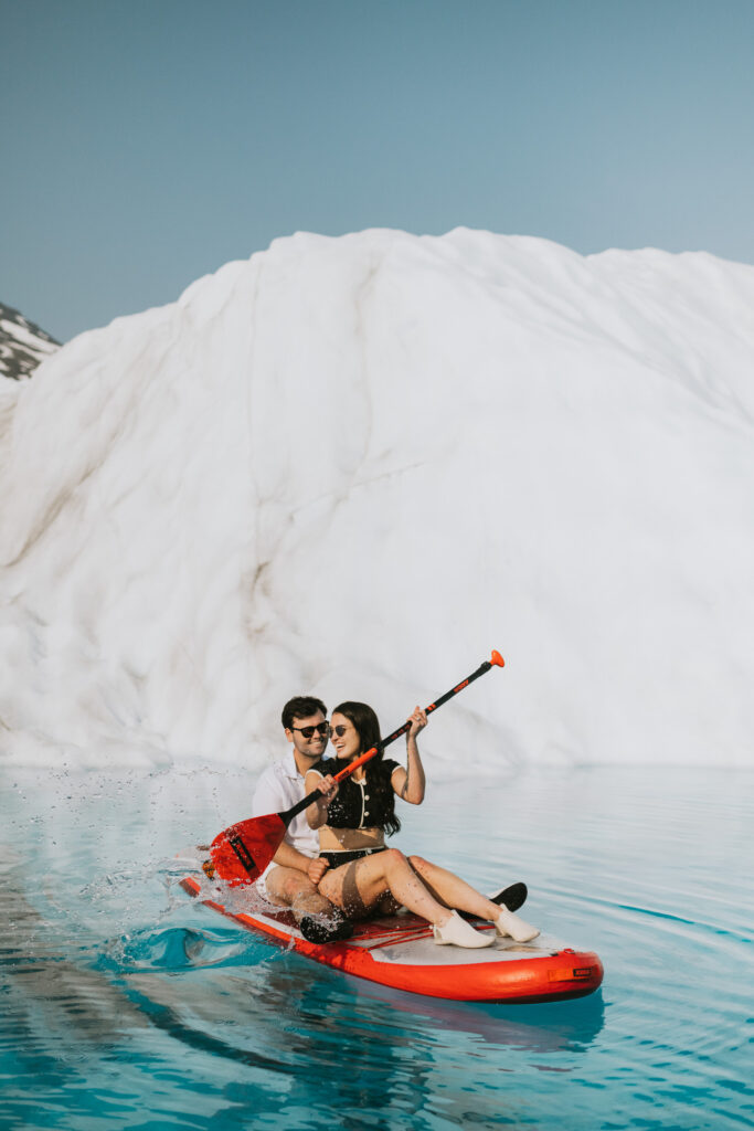 A couple paddle boarding on a glacier lake smiling delightedly on a sunny day for their engagement photo.