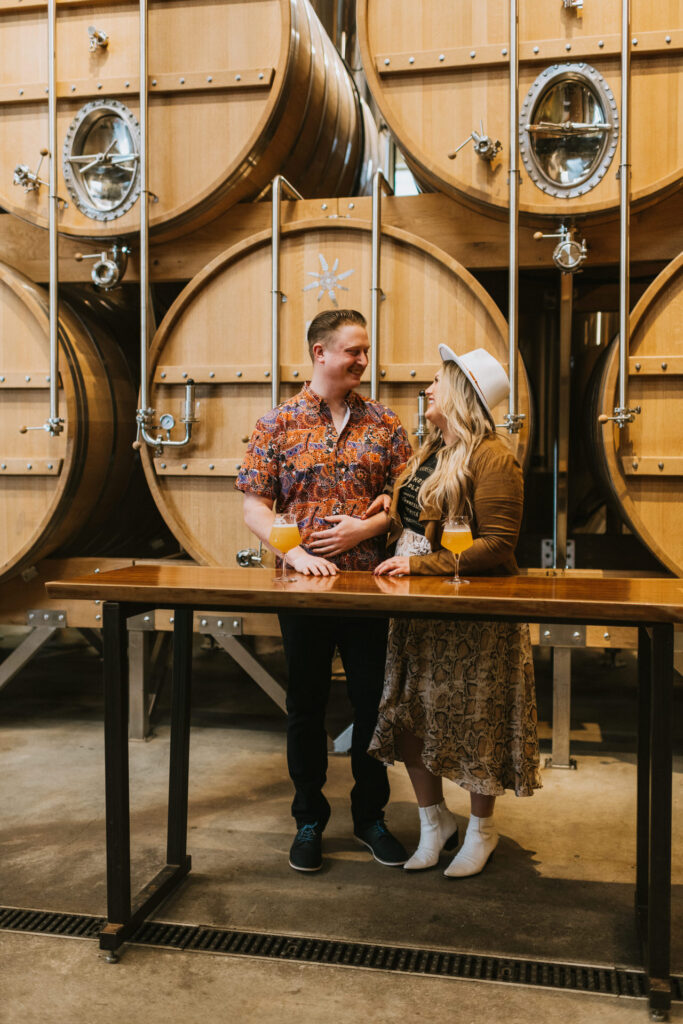 A couple standing and smiling at each other, holding beers in a brewery with large wooden barrels in the background during their engagement photo session.
