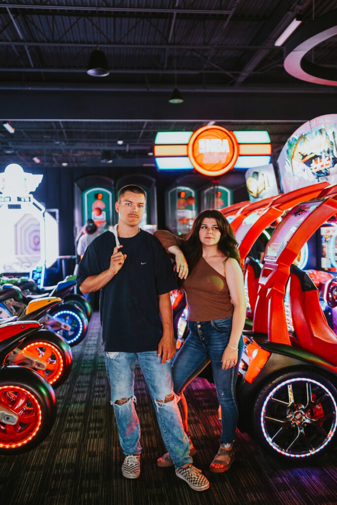 A young man and woman standing in an arcade, surrounded by colorful racing game machines, posing for their engagement photos.