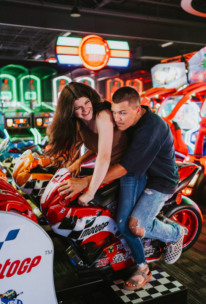 A couple playing on an arcade motorcycle racing game, laughing joyfully in a colorful game room setting during their engagement photo shoot.