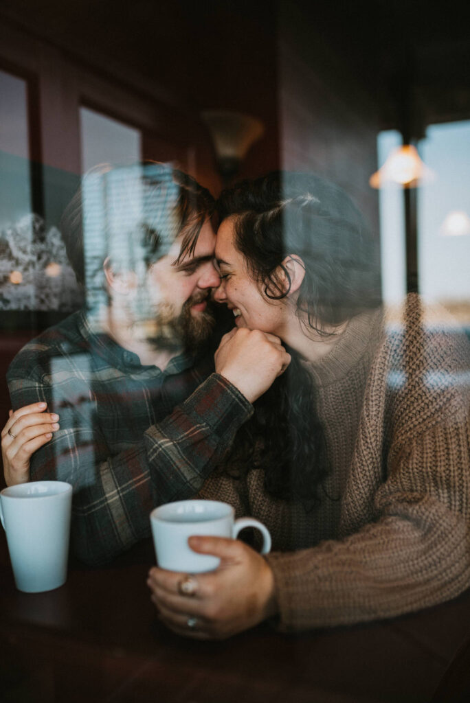 A couple holding mugs and smiling at each other affectionately through a window, with soft reflections visible on the glass, perfectly capturing engagement photo ideas.