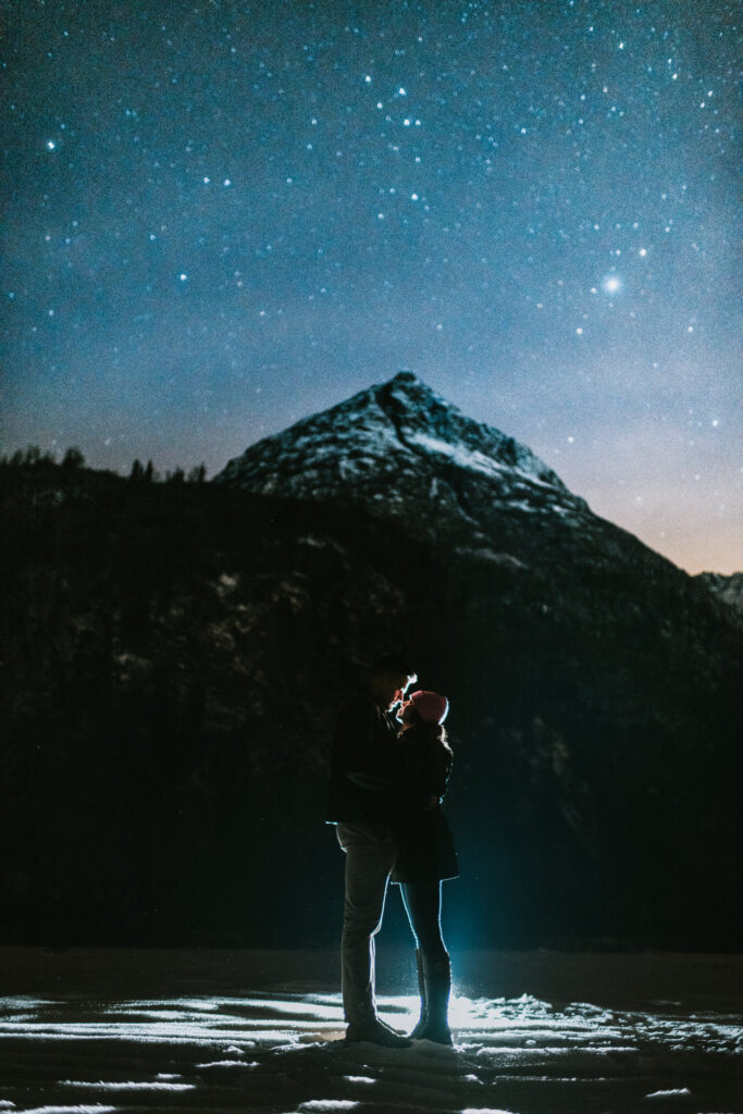 A couple embraces under a starry sky with a snow-capped mountain in the background, capturing an ideal moment for engagement photos.
