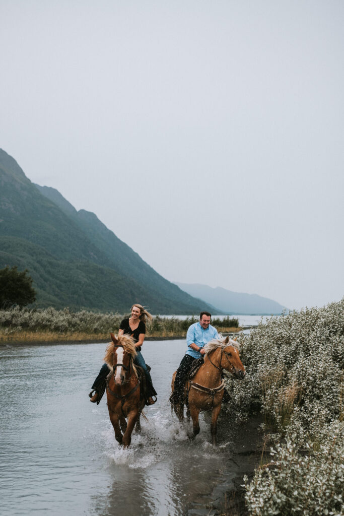 Two people joyfully riding horses through a shallow river with mountains in the background for their engagement photos.