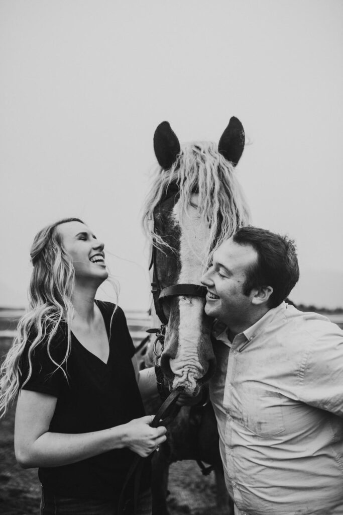 A joyful couple laughing and interacting with a horse in a foggy field during their engagement photo session.