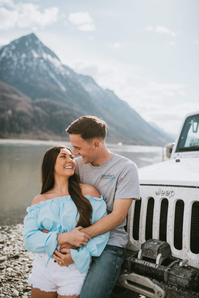 A happy couple embracing beside a white jeep for their engagement photos, with a mountainous backdrop and a cloudy sky.