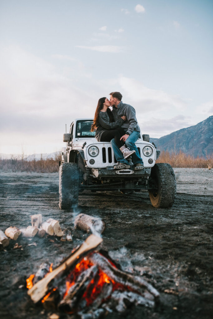 A couple kissing on the hood of a white off-road vehicle near a campfire, with mountains in the background, captured by an engagement photographer.