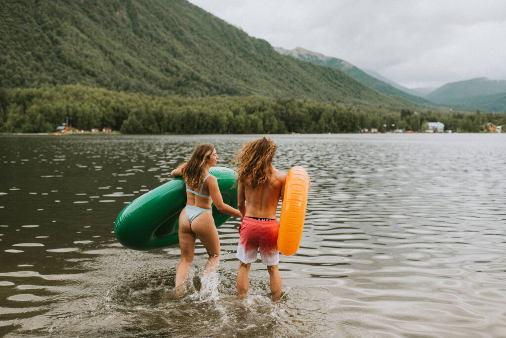 Two people holding pool floats wade into a lake for their engagement photos, with mountainous scenery in the background.