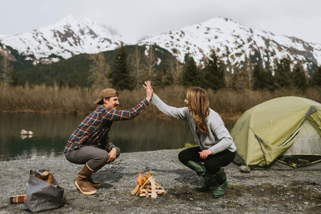 A man and a woman high-fiving by a campfire near a tent with a scenic mountain backdrop during their engagement photo shoot.